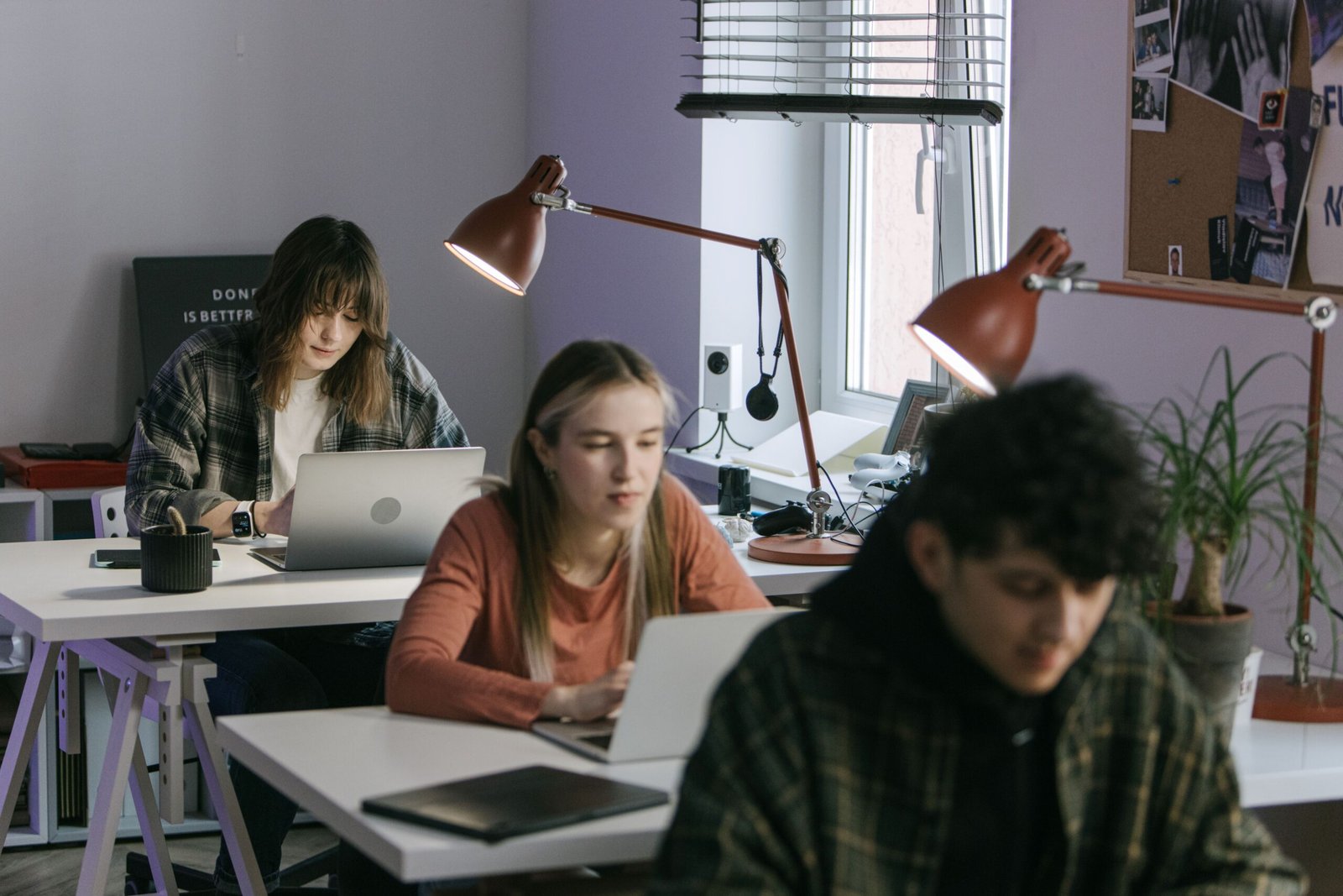 Group of students working at desks with laptops in a bright classroom/study area.