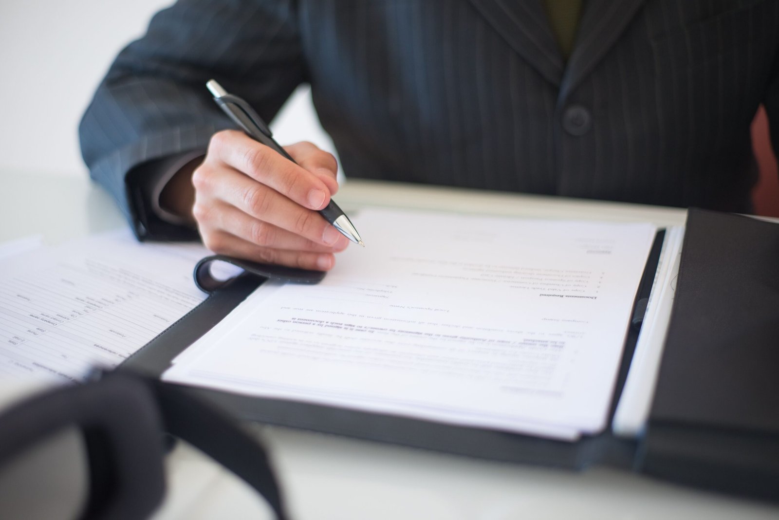 Businessperson's hand writes with a pen over documents in a binder on a desk, glasses at the edge of the papers