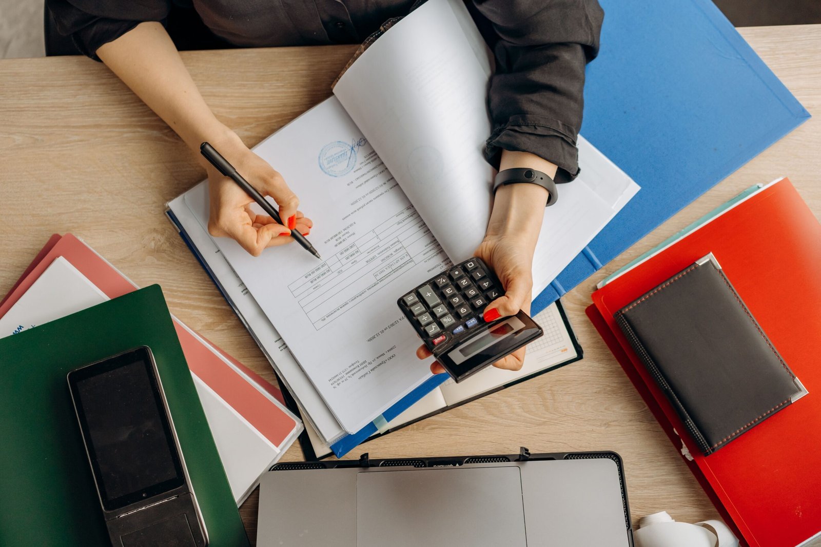 Person using a calculator and writing on documents at a cluttered desk with notebooks and folders nearby