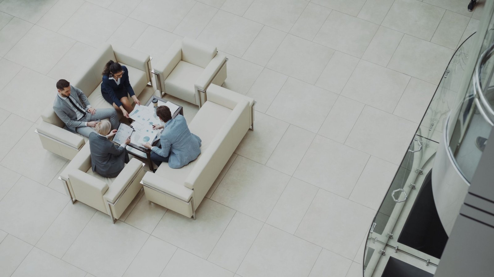 Top-down view of four professionals in a modern lobby seated around a glass table reviewing charts and documents in a bright atrium.